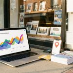 Lancer une offre localement : Google Ads, référencement ou terrain ? Vitrine d’un commerce de quartier accompagnée d’un ordinateur affichant des formes analytiques abstraites, d’un smartphone avec une épingle de localisation et de supports promotionnels vierges.